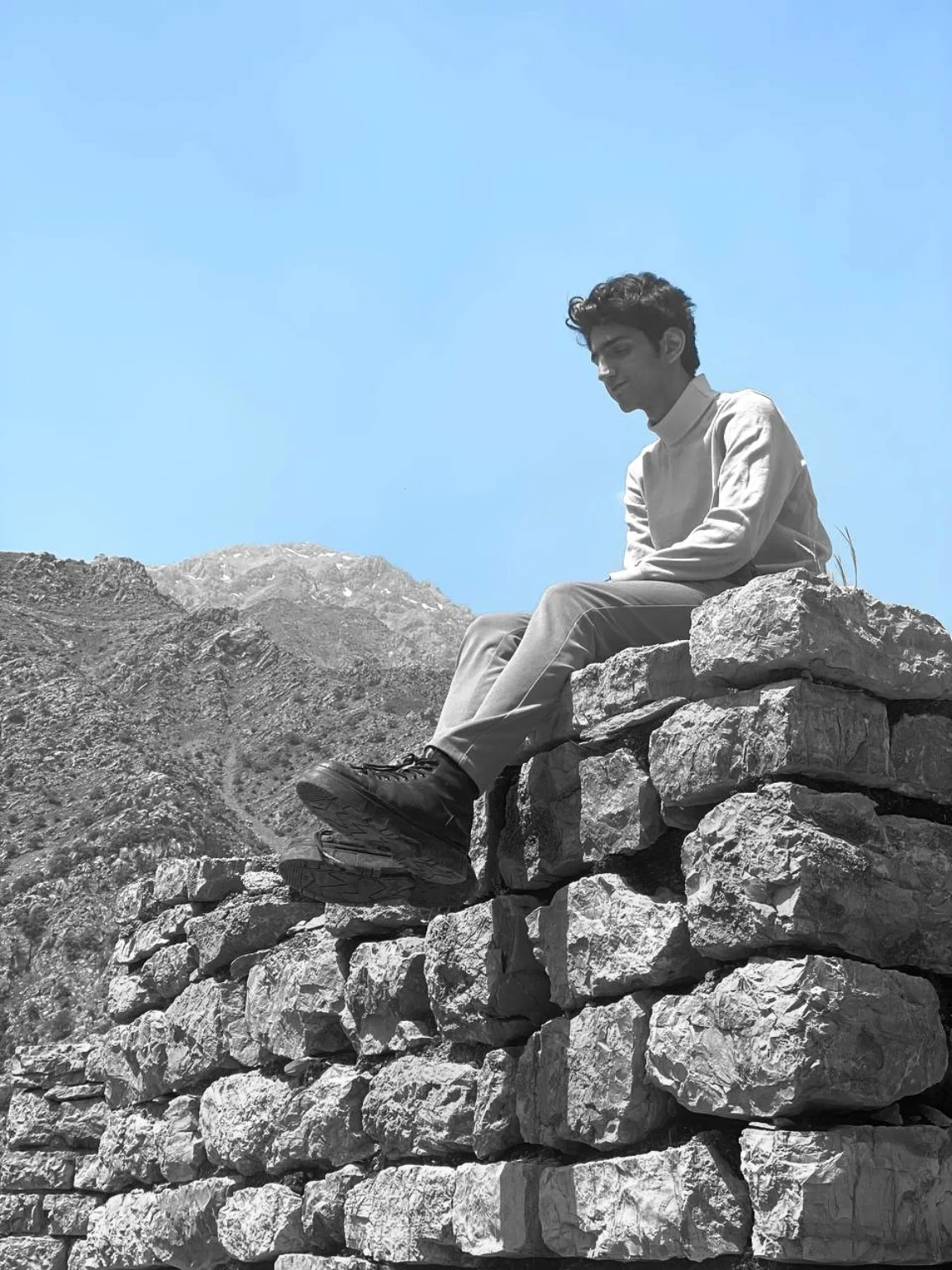Kiarash seated on a stone wall in a mountain landscape under a blue sky