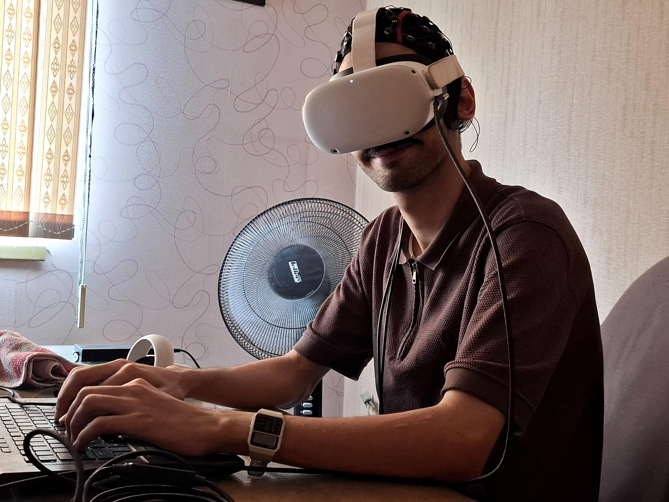 Kiarash wearing an EEG cap and VR headset while working at a laptop, August 2026